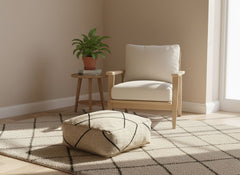 Beige Berber pouf on a neutral rug next to a wooden chair and plant