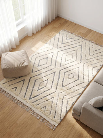 Soft ivory geometric rug with grey lines viewed from above beside a beige sofa and pouf in a sunlit living space.