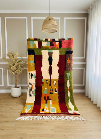 Handwoven Moroccan rug displayed vertically showing bold abstract shapes in red, mustard, green, and cream tones.