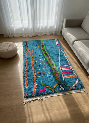 Small Moroccan wool rug in turquoise with colorful details placed beside a sofa in a sunlit corner.