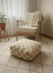 “Beige wool checkered pouffe beside armchair in cozy room.”