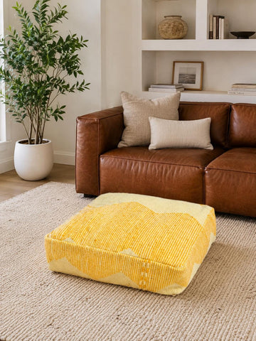 Mustard Moroccan floor pouf styled on a neutral rug in a modern living room with a brown leather sofa.